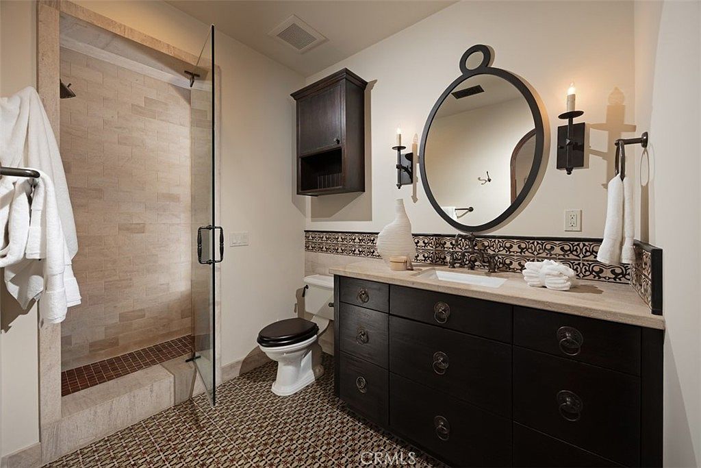 This is a bathroom featuring a dark wood vanity with a light countertop, a decorative backsplash, and a round mirror with sconces. A toilet is positioned next to the vanity, and a glass-enclosed shower is visible to the left. The floor is tiled with a patterned design, creating a warm and inviting atmosphere.