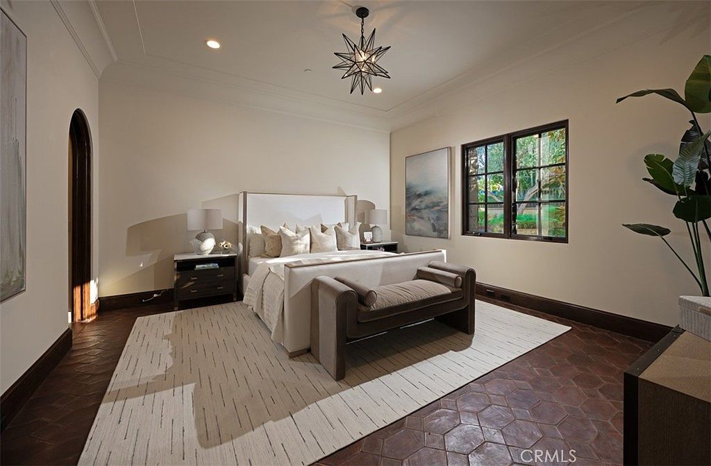 This is a primary bedroom featuring a large bed with a white headboard and neutral-toned bedding. The room has a warm and inviting atmosphere, with natural light streaming in through a window. The flooring is a unique tile pattern, and a stylish bench sits at the foot of the bed, adding to the room's elegance.