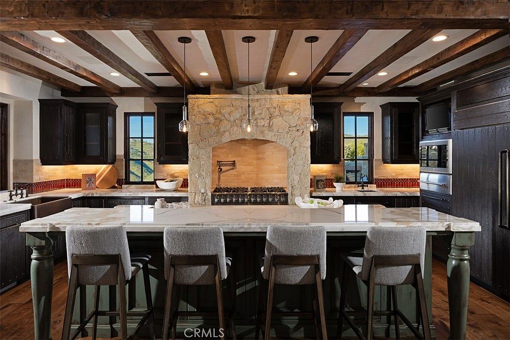 This is a well-appointed kitchen featuring dark wood cabinetry, a stone range hood, and a large island with seating. The ceiling has exposed wooden beams, and pendant lights hang above the island. The overall style is rustic and luxurious, creating a warm and inviting atmosphere.