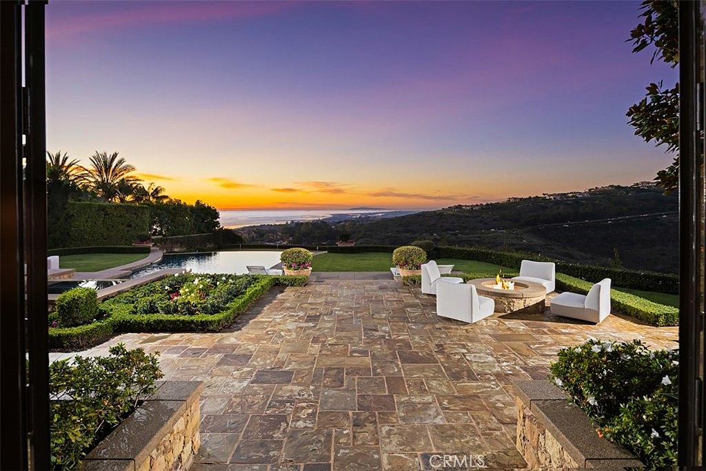 This image showcases a luxurious outdoor patio area at sunset. The patio features stone flooring, a fire pit surrounded by comfortable white seating, and meticulously manicured landscaping. In the background, there's a swimming pool and a stunning view of the horizon, suggesting a high-end property with exceptional outdoor living spaces.