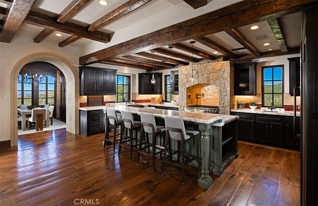 This is a spacious kitchen featuring dark wood cabinetry, a large central island with seating, and a stone-clad range hood. The kitchen has exposed wooden beams on the ceiling and hardwood floors, creating a rustic yet luxurious feel. The open layout connects to a dining area visible through an arched doorway.