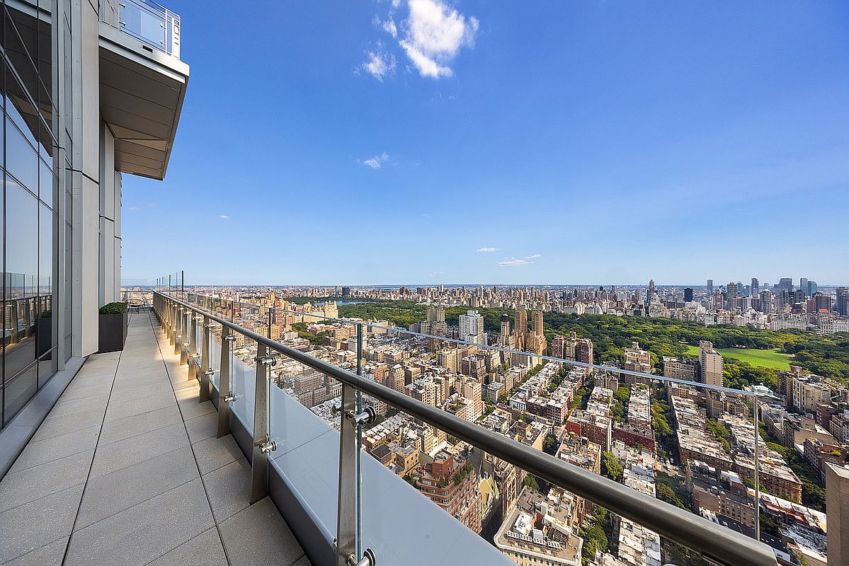 This high-rise balcony offers a breathtaking, panoramic view of Central Park and the surrounding New York City skyline. The space features sleek, modern glass railings and light-colored stone flooring, creating a sophisticated and airy outdoor living area. The perspective captures the immense scale of the city from a luxurious vantage point, emphasizing the exclusivity and prime location of the property.