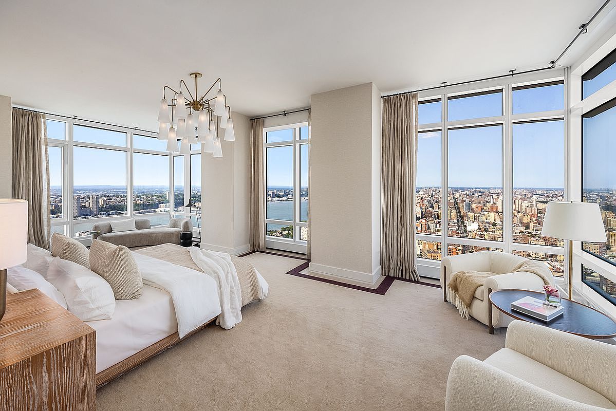 This luxurious primary bedroom features floor-to-ceiling windows that offer breathtaking, panoramic views of the city skyline and waterfront. The room is elegantly furnished with a plush king-sized bed, a cozy seating area with two armchairs, and a sophisticated chandelier that serves as a focal point. The neutral color palette and soft carpeting create a serene, high-end atmosphere perfect for a premium urban residence.
