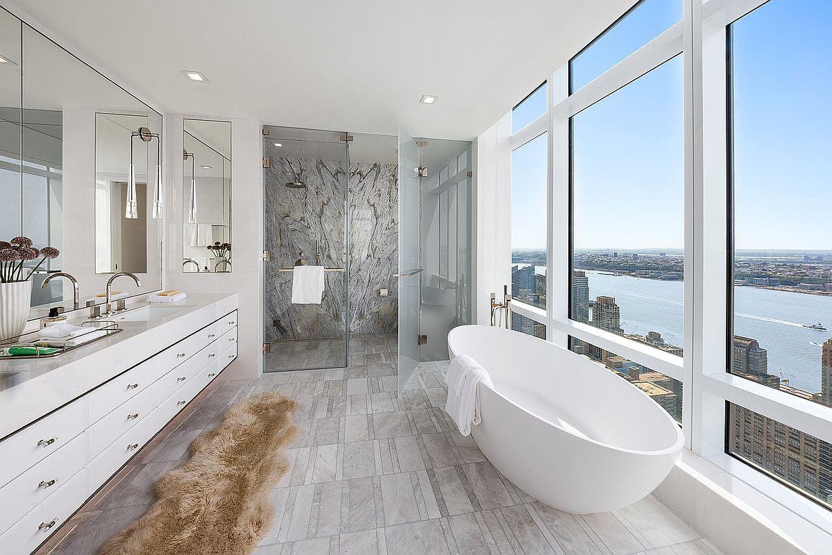 This luxurious primary bathroom features a sleek, modern design with a freestanding soaking tub positioned next to floor-to-ceiling windows that offer a breathtaking panoramic view of the city skyline and river. The space includes a long, white double vanity with polished fixtures, a spacious walk-in shower with striking marble-patterned tile, and elegant light-toned stone flooring accented by a plush faux-fur rug. The overall aesthetic is sophisticated, airy, and high-end, emphasizing the dramatic urban vista.