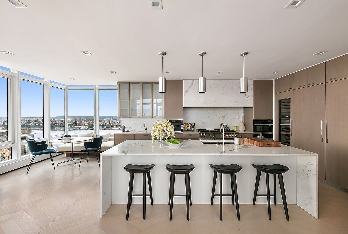 This sophisticated, modern kitchen features a large waterfall-edge marble island with four sleek black stools, serving as the centerpiece of the open-concept space. The room is bathed in natural light from floor-to-ceiling windows that offer a panoramic city view, complemented by high-end cabinetry and professional-grade appliances. The clean lines, neutral color palette, and minimalist aesthetic create an atmosphere of luxury and refined urban living.