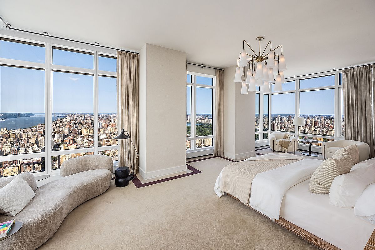This luxurious primary bedroom features floor-to-ceiling windows that offer breathtaking, panoramic views of the city skyline and river. The room is elegantly furnished with a plush, curved chaise lounge, a modern bed with neutral-toned bedding, and a sophisticated, multi-tiered chandelier that serves as a focal point. The overall aesthetic is one of refined, high-end urban living, characterized by soft textures, a neutral color palette, and an expansive, airy atmosphere.