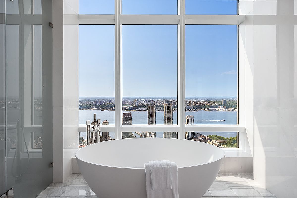 This luxurious primary bathroom features a sleek, white freestanding soaking tub positioned perfectly in front of floor-to-ceiling windows that offer a breathtaking, panoramic view of the river and city skyline. The minimalist design is complemented by polished marble flooring and clean, white tiled walls, creating an atmosphere of sophisticated tranquility. The perspective is centered on the tub, emphasizing the seamless integration of high-end interior design with the dramatic urban landscape outside.