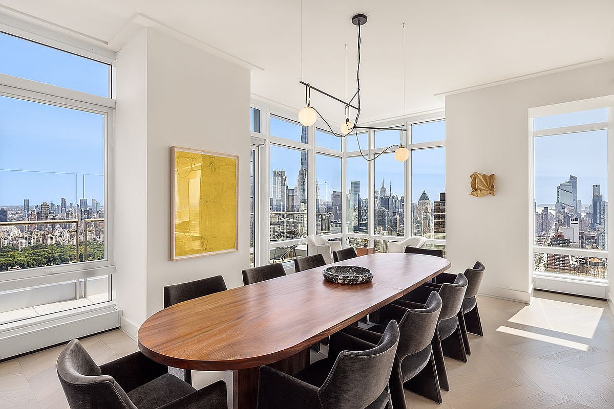 This sophisticated dining room features a large, oval-shaped wooden table surrounded by dark, upholstered chairs, set against a backdrop of floor-to-ceiling windows that offer a breathtaking panoramic view of the New York City skyline and Central Park. A modern, minimalist chandelier hangs above the table, while a vibrant yellow abstract painting adds a pop of color to the clean, white-walled space. The room exudes a sense of luxury and high-end urban living, with light-toned herringbone flooring completing the elegant aesthetic.
