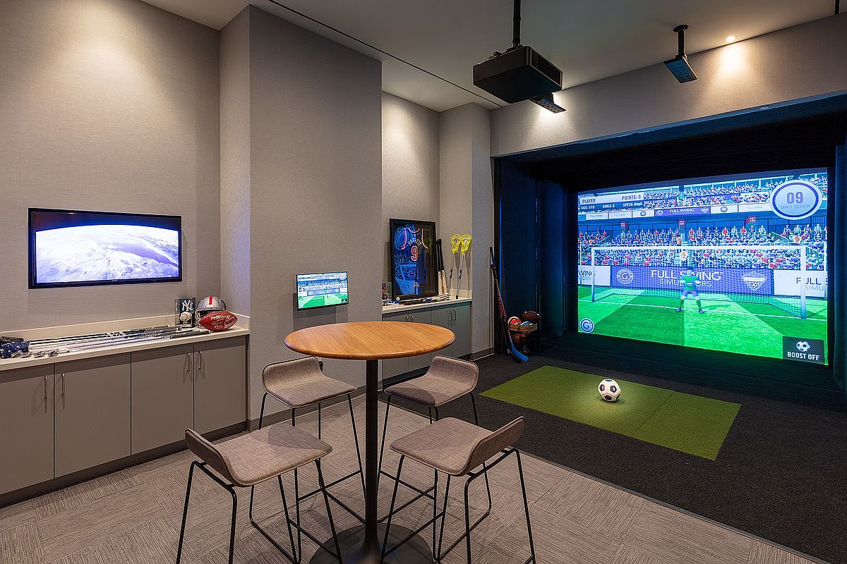 This high-end indoor sports simulator room features a large projection screen displaying a soccer game, complete with a turf mat and a soccer ball ready for play. The space is furnished with a modern circular high-top table and four chairs, alongside built-in cabinetry displaying sports memorabilia and golf clubs. The room offers a sophisticated, recreational atmosphere perfect for entertainment and leisure.