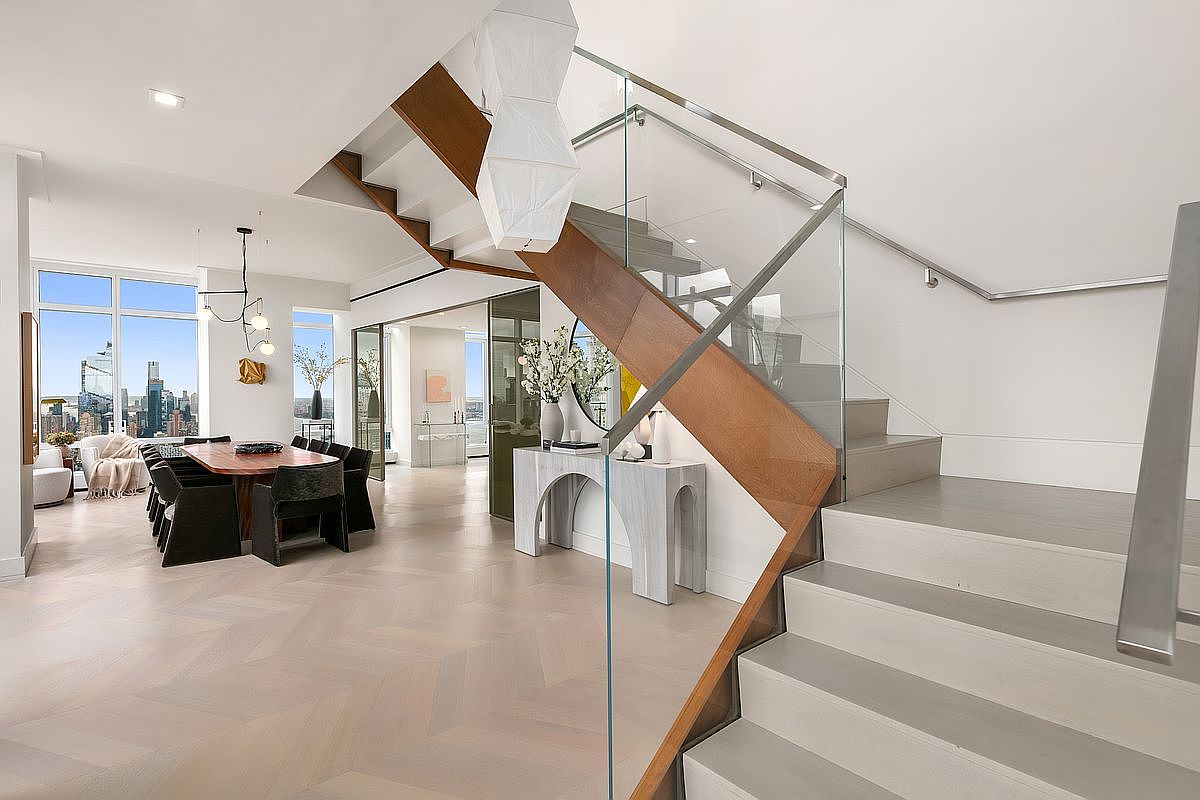This sophisticated interior space features a striking floating staircase with light wood treads and a sleek glass railing, serving as a central architectural focal point. To the left, an elegant dining area is visible, complete with a large wooden table and floor-to-ceiling windows that offer a panoramic city view. The overall aesthetic is modern and luxurious, characterized by clean lines, light-toned herringbone flooring, and a bright, airy atmosphere.
