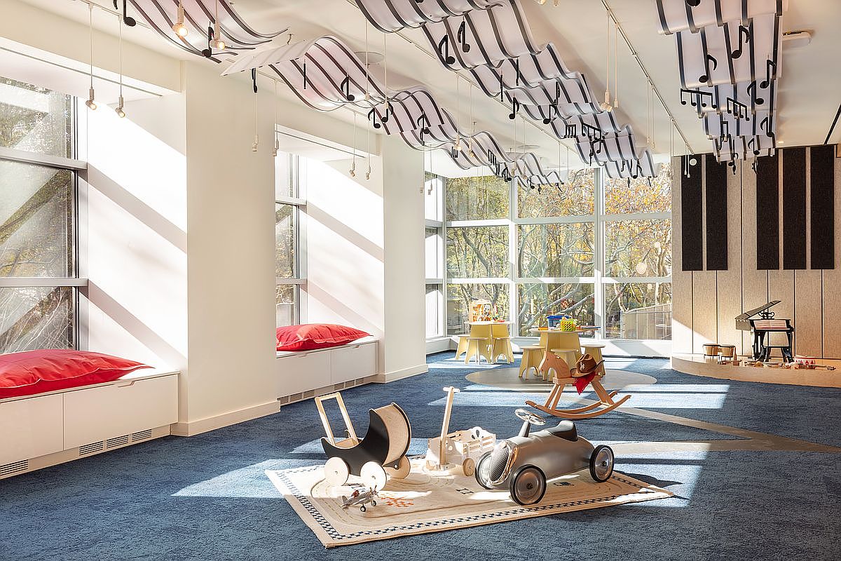 This vibrant children's playroom features a whimsical musical theme with staff-notation ribbons suspended from the ceiling and a dedicated stage area with a miniature piano. The space is equipped with various toys, including a rocking horse and a vintage-style ride-on car, set against a backdrop of large windows that flood the room with natural light. The blue carpeting and built-in window seating create a cozy, inviting atmosphere perfect for a high-end residential amenity space.