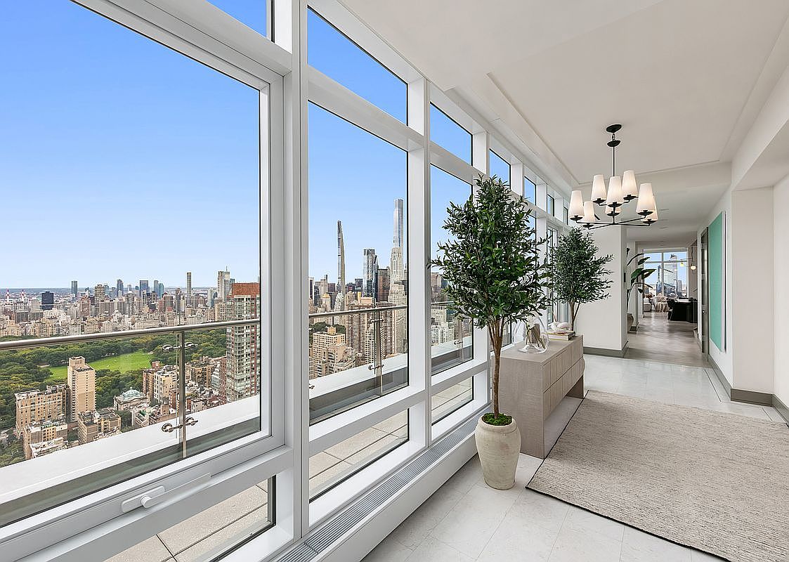 This elegant, sun-drenched hallway features floor-to-ceiling windows that offer a breathtaking, panoramic view of Central Park and the New York City skyline. The space is minimalist and sophisticated, accented by two potted olive trees, a sleek console table, and a modern chandelier that adds a touch of refined luxury. The perspective captures the seamless transition between the bright, airy interior and the expansive urban landscape outside.