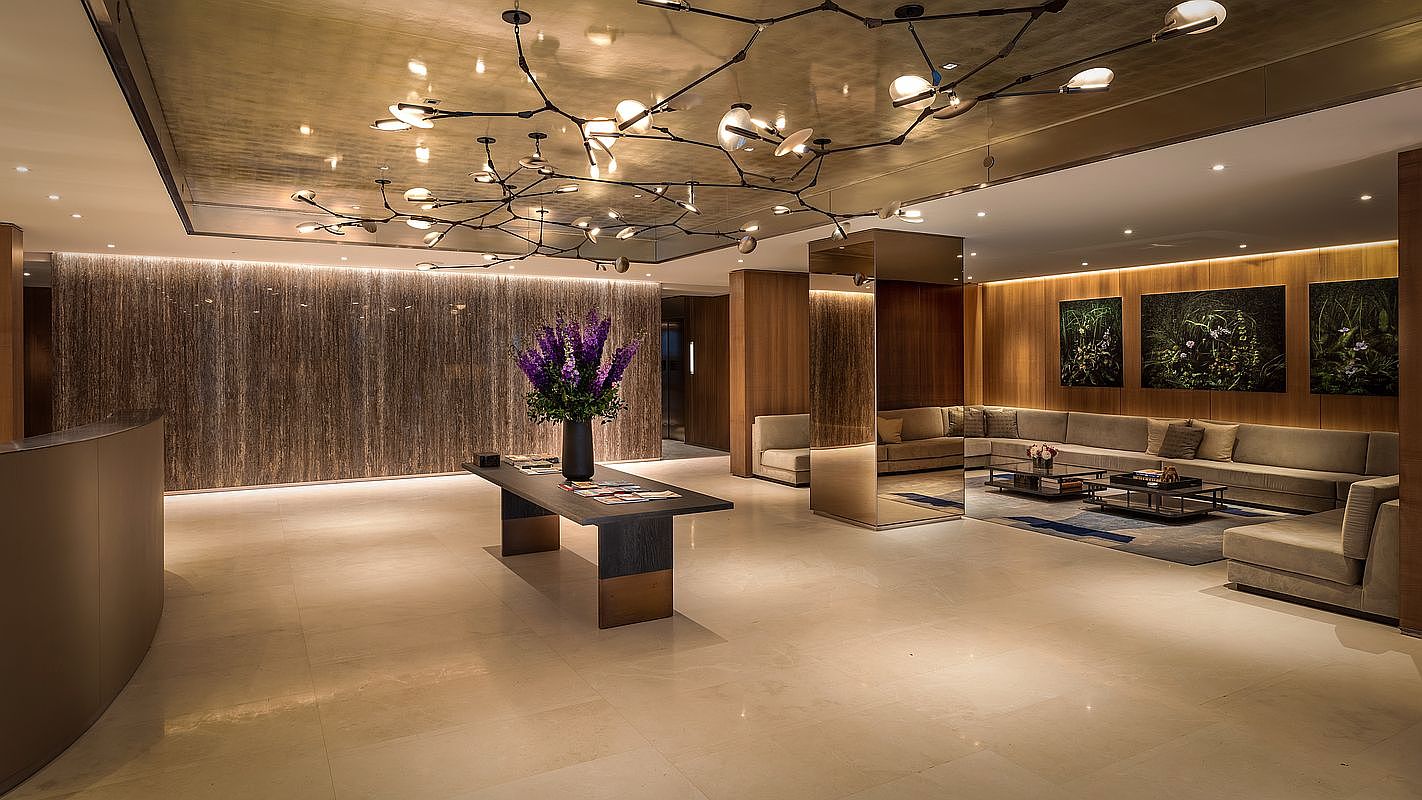 This sophisticated lobby or lounge area features a high-end, contemporary design with a striking branching chandelier as the focal point. The space is anchored by a sleek central console table with a vibrant floral arrangement, complemented by a large, plush sectional sofa area and warm wood-paneled walls. The perspective is a wide, eye-level shot that captures the luxurious, hotel-like atmosphere and the seamless integration of ambient lighting and textured wall treatments.