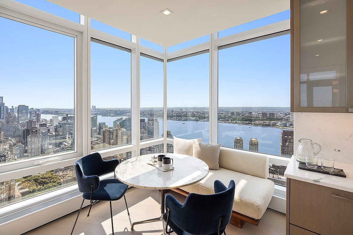 This sophisticated breakfast nook features a circular marble-topped table paired with two navy blue upholstered armchairs and a plush, cream-colored bench seat. The space is defined by floor-to-ceiling corner windows that offer a breathtaking, panoramic view of the city skyline and river below. The modern, minimalist aesthetic creates an airy and luxurious atmosphere, perfect for enjoying morning coffee while overlooking the urban landscape.