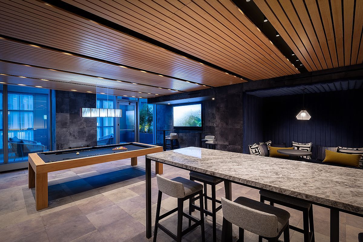 This sophisticated communal lounge area features a sleek pool table as a centerpiece, complemented by a high-top marble bar with modern seating. The space is defined by a striking wood-slat ceiling, dark stone accent walls, and a cozy recessed seating nook with plush cushions. The overall aesthetic is upscale and contemporary, designed for social interaction and relaxation in a luxury residential setting.