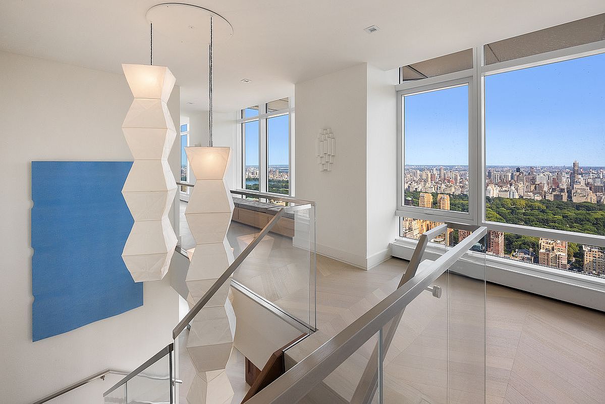 This sophisticated interior space features a modern staircase with a sleek glass railing, leading to an upper landing bathed in natural light. The area is accented by striking, sculptural pendant lights and a large blue abstract art piece, while floor-to-ceiling windows offer a breathtaking, expansive view of Central Park and the New York City skyline. The minimalist design and light-toned hardwood flooring create an airy, luxurious atmosphere perfect for a high-end urban residence.