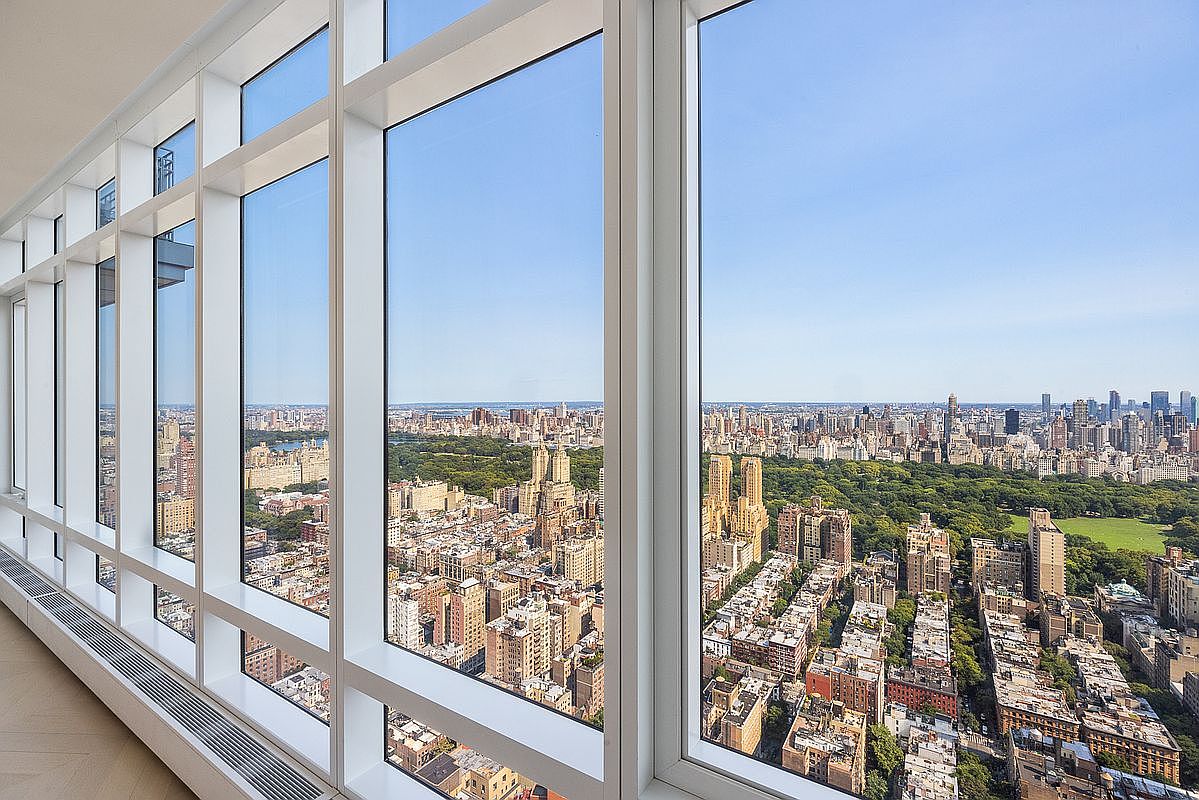 This image captures a high-rise interior space defined by floor-to-ceiling windows that offer a breathtaking, panoramic aerial view of Central Park and the surrounding New York City skyline. The minimalist white window frames create a clean, modern aesthetic that emphasizes the expansive urban vista. The perspective is from inside a luxury apartment, highlighting the seamless connection between the sophisticated interior architecture and the iconic cityscape outside.