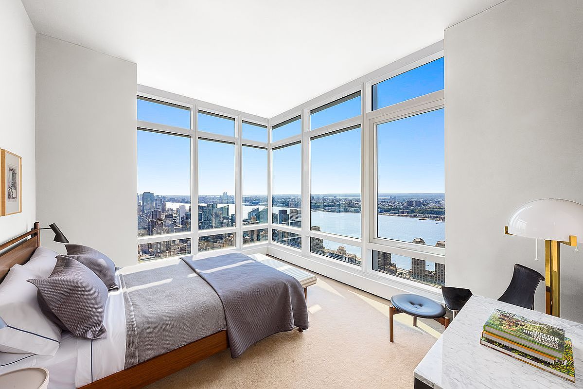 This modern, high-rise primary bedroom features floor-to-ceiling corner windows that offer a breathtaking, panoramic view of the city skyline and river. The room is furnished with a sleek, low-profile wooden bed frame, a marble-topped desk, and minimalist decor, creating a sophisticated and serene atmosphere. The perspective is captured from a slightly elevated angle, emphasizing the expansive glass walls and the luxurious, airy feel of the space.