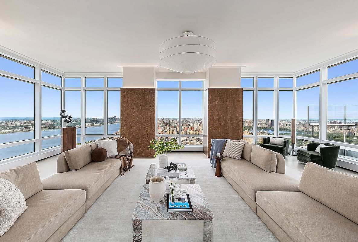 This expansive, high-end living room features a symmetrical layout with two large, plush beige sofas facing each other across a pair of elegant marble coffee tables. Floor-to-ceiling windows wrap around the space, offering breathtaking, panoramic views of the city skyline and river, while warm wood-paneled columns add a touch of natural sophistication. The room is anchored by a modern, tiered pendant light fixture, creating a bright, airy, and luxurious atmosphere perfect for high-rise urban living.