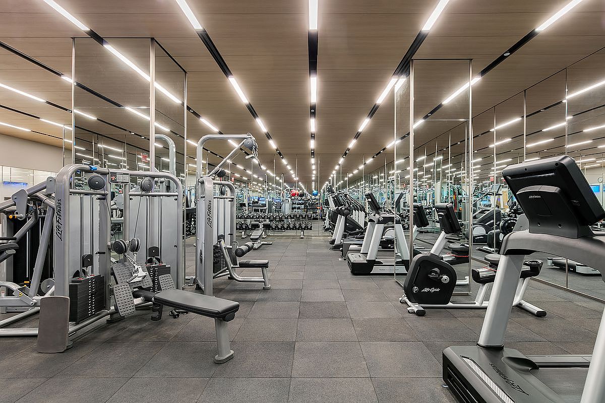 This professional-grade home gym features a sleek, modern design with mirrored walls that create an expansive, infinite perspective. The space is equipped with high-end Life Fitness strength training machines and treadmills, all set upon durable, dark rubberized flooring. Linear recessed ceiling lighting provides a bright, clean aesthetic, making the facility feel both sophisticated and highly functional for residents.