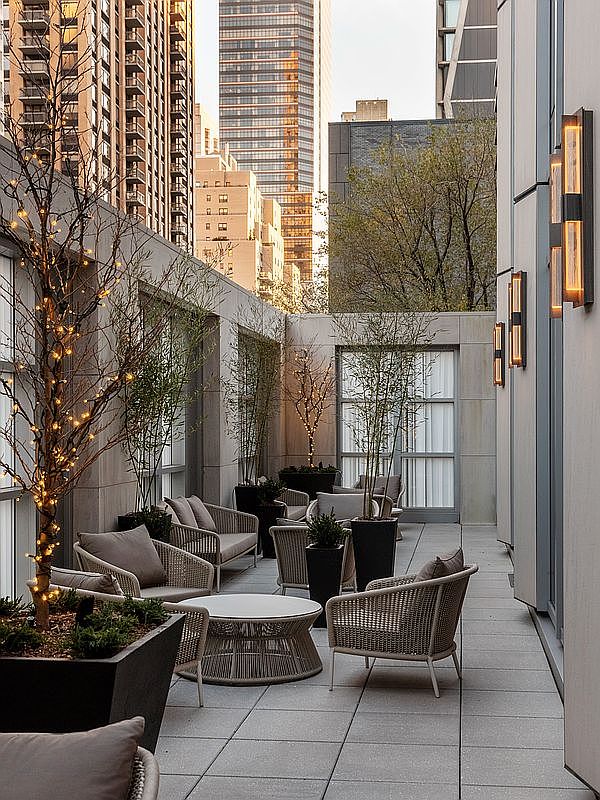 This sophisticated urban terrace offers a serene outdoor retreat, featuring modern woven patio furniture arranged for comfortable seating. The space is adorned with potted greenery and a tree wrapped in warm fairy lights, creating an inviting atmosphere against the backdrop of towering city skyscrapers. The clean lines of the stone flooring and architectural wall lighting enhance the contemporary, high-end aesthetic of this private balcony.