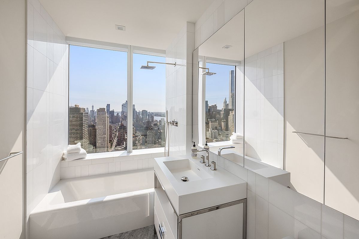 This luxurious, modern bathroom features a sleek, minimalist design with a deep soaking tub positioned directly against a floor-to-ceiling window that offers a breathtaking, panoramic view of the city skyline. The space is anchored by a contemporary white vanity with a rectangular sink and polished chrome fixtures, complemented by large-format white wall tiles and a mirrored cabinet. The bright, airy atmosphere creates a sophisticated spa-like retreat high above the urban landscape.