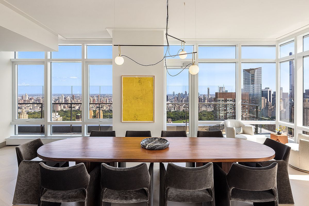 This sophisticated dining room features a large, oval wooden table surrounded by plush, dark grey velvet chairs, centered beneath a modern, minimalist chandelier. The room is defined by floor-to-ceiling windows that offer a breathtaking, panoramic view of the city skyline and Central Park. A vibrant yellow abstract painting serves as a bold focal point on the central pillar, enhancing the room's luxurious and contemporary aesthetic.