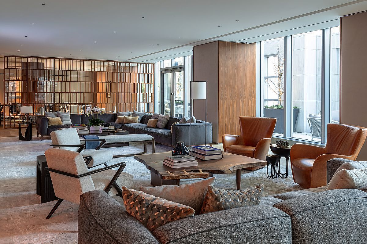 This sophisticated and spacious lounge area features a contemporary design with a mix of plush grey sectional sofas, warm leather armchairs, and a unique live-edge wood coffee table. The room is anchored by a large, textured area rug and illuminated by floor-to-ceiling windows that provide ample natural light and a view of an outdoor terrace. A decorative wooden screen adds architectural interest, creating a refined and inviting atmosphere suitable for a high-end residential or hotel lobby setting.