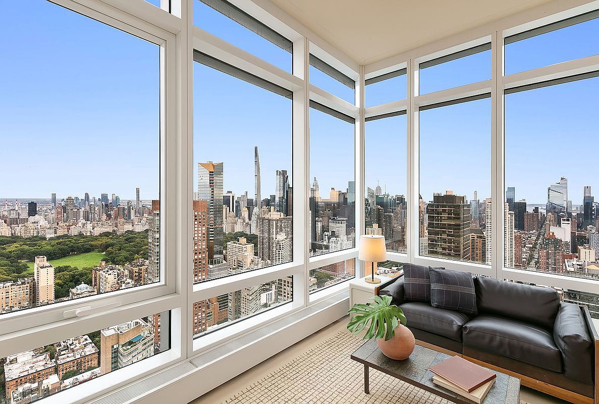 This high-rise living room features floor-to-ceiling corner windows that offer a breathtaking, panoramic view of the New York City skyline and Central Park. The space is furnished with a sleek, dark leather sofa, a minimalist coffee table, and a potted plant, creating a sophisticated and modern urban retreat. The perspective captures the seamless blend of luxury interior design with the dramatic, expansive cityscape outside.