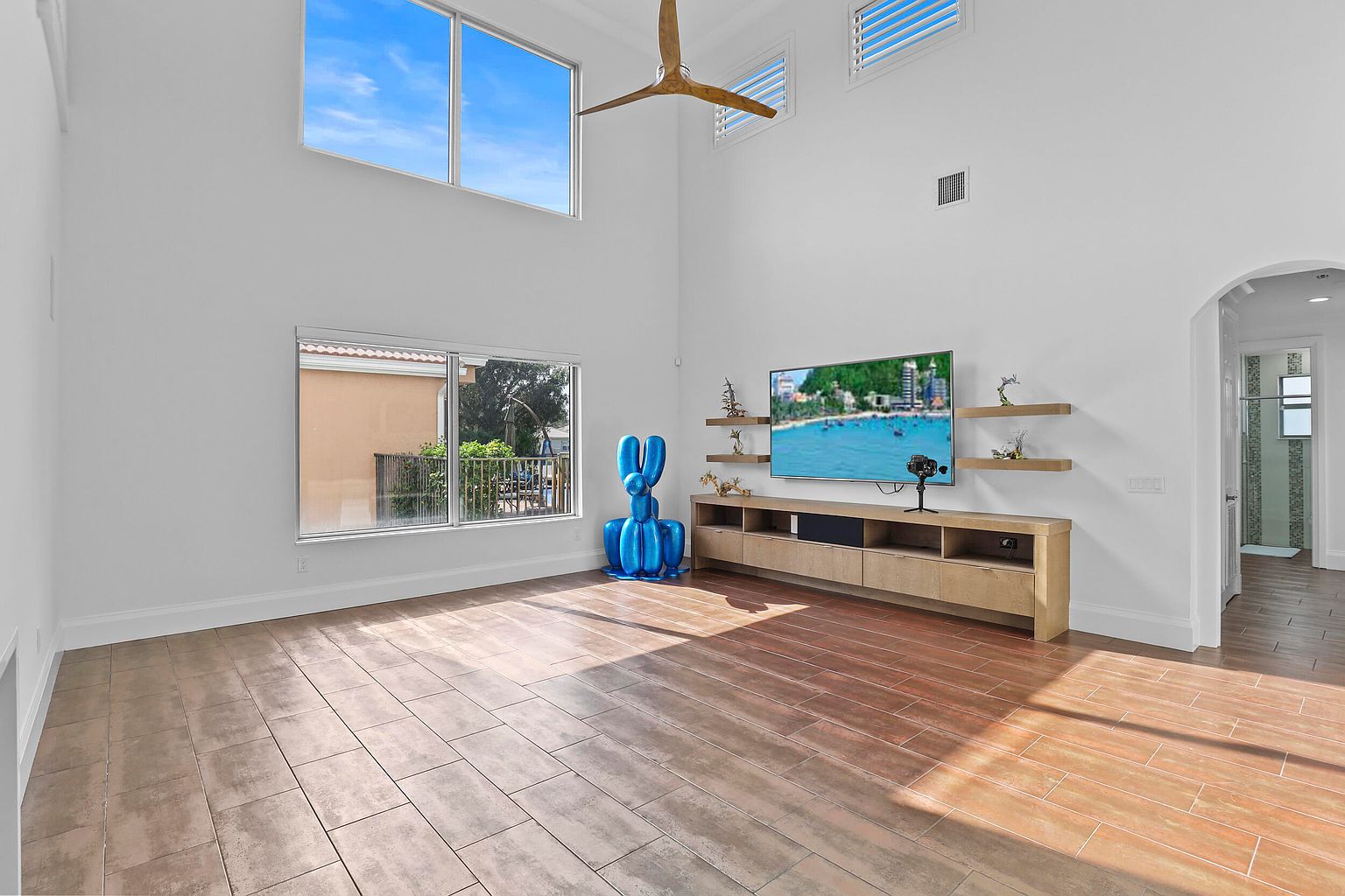 This is an interior shot of a living room featuring high ceilings and a modern aesthetic. The room is well-lit with natural light coming through large windows. A large television is mounted above a long, light-colored media console, and a blue balloon animal sculpture adds a pop of color. The flooring is tiled and the walls are painted white, contributing to a bright and spacious feel.