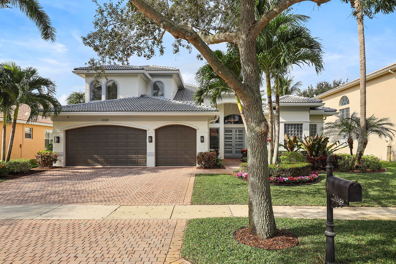 This is a front exterior view of a two-story home with a well-manicured lawn and mature landscaping. The house features a gray tile roof, a double garage with brown doors, and a light-colored facade. A brick driveway leads to the garage, and palm trees and other greenery add to the curb appeal, creating an inviting and luxurious impression.
