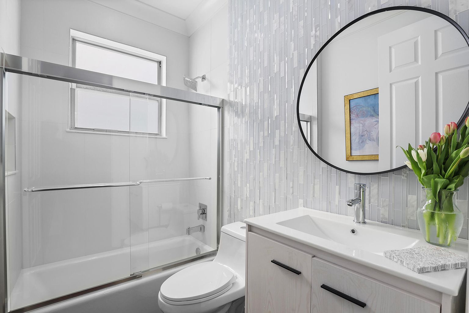 This is a well-lit bathroom featuring a bathtub with a glass enclosure, a white toilet, and a vanity with a white countertop and dark hardware. The wall behind the vanity is adorned with vertical mosaic tiles, and a round mirror hangs above the sink. The overall impression is clean, modern, and stylish.