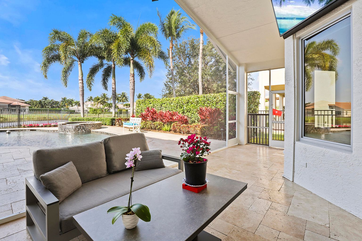 This is a beautifully staged outdoor patio area, perfect for relaxation and entertaining. The space features a comfortable gray sofa and a modern coffee table adorned with potted plants. The patio overlooks a stunning pool area with lush landscaping, including palm trees and vibrant red foliage, creating a serene and inviting atmosphere.