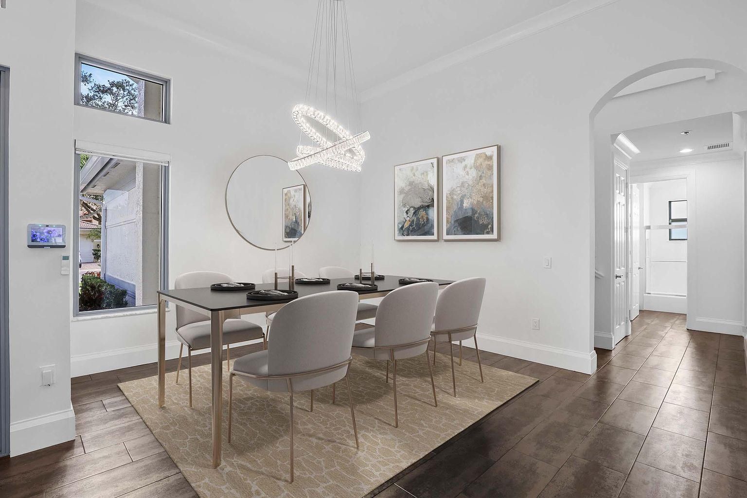 This is an interior shot of a dining room featuring a modern aesthetic. A black dining table with light-colored chairs sits atop a patterned rug, illuminated by a contemporary chandelier. The room is decorated with abstract art and a round mirror, enhancing the bright and airy feel, while the dark wood flooring adds a touch of warmth.