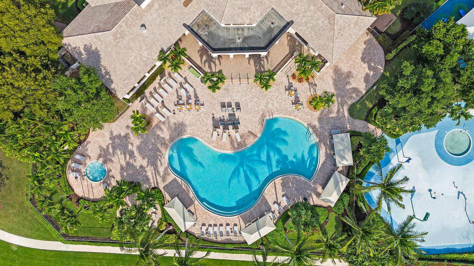 This aerial shot showcases a luxurious pool and spa area, surrounded by lush landscaping and elegant lounge furniture. The pool's unique shape and clear blue water create a focal point, complemented by the surrounding palm trees and manicured grounds. The overall impression is one of relaxation and upscale resort living.