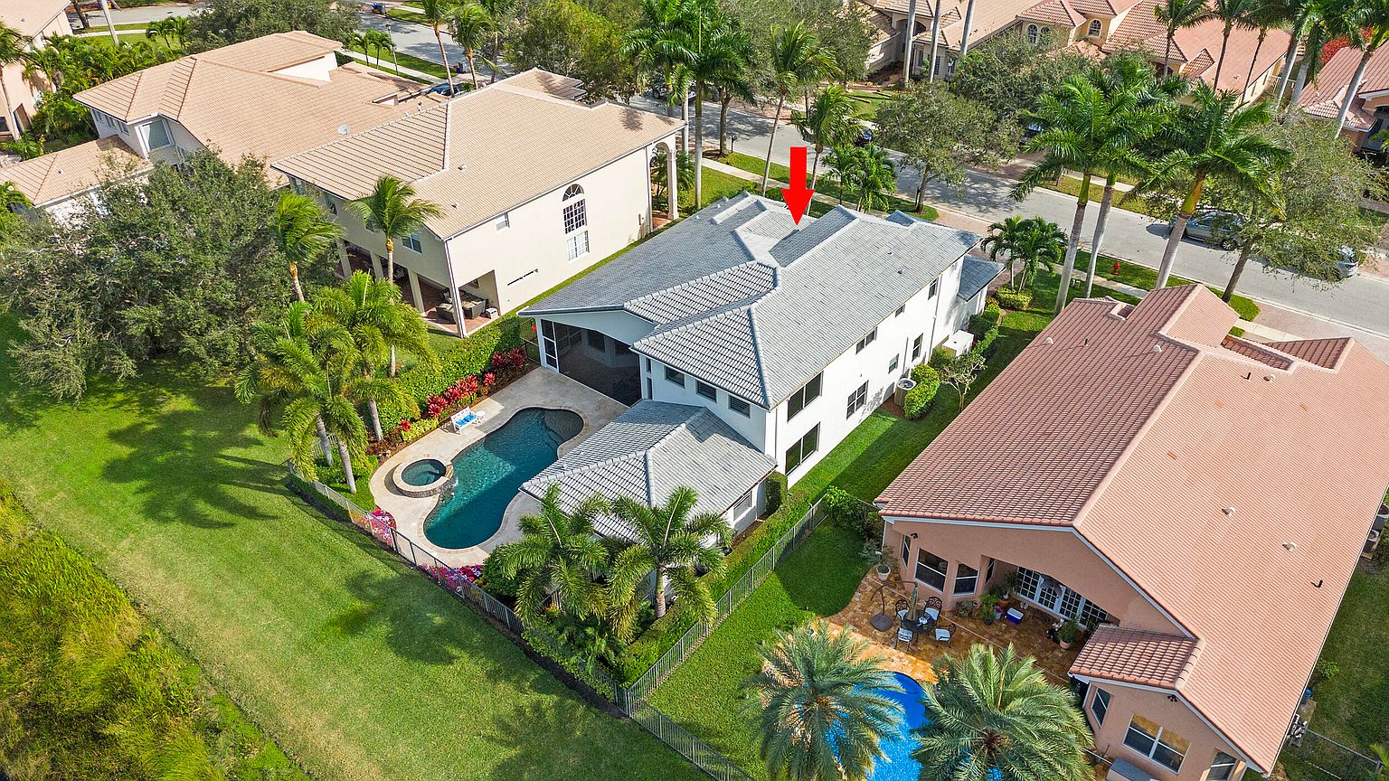 This aerial view showcases a well-maintained two-story home with a gray tile roof, a private pool and spa surrounded by lush landscaping, and a fenced backyard. The property is situated in a residential neighborhood with mature trees and manicured lawns, offering a sense of privacy and luxury. The perspective is from above, looking down at a slight angle, highlighting the home's layout and outdoor amenities.