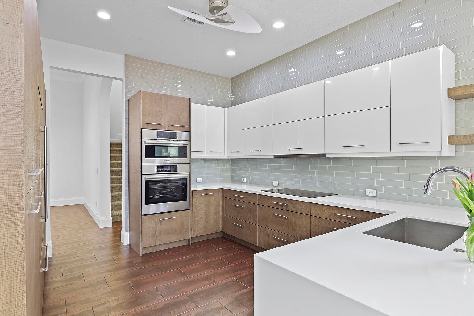 This is a modern kitchen with a combination of white and wood-grain cabinets, stainless steel appliances, and a white countertop. The backsplash is a light gray subway tile, and the flooring is a dark wood-look tile. The kitchen features a built-in oven and microwave, and a sleek cooktop on the counter.