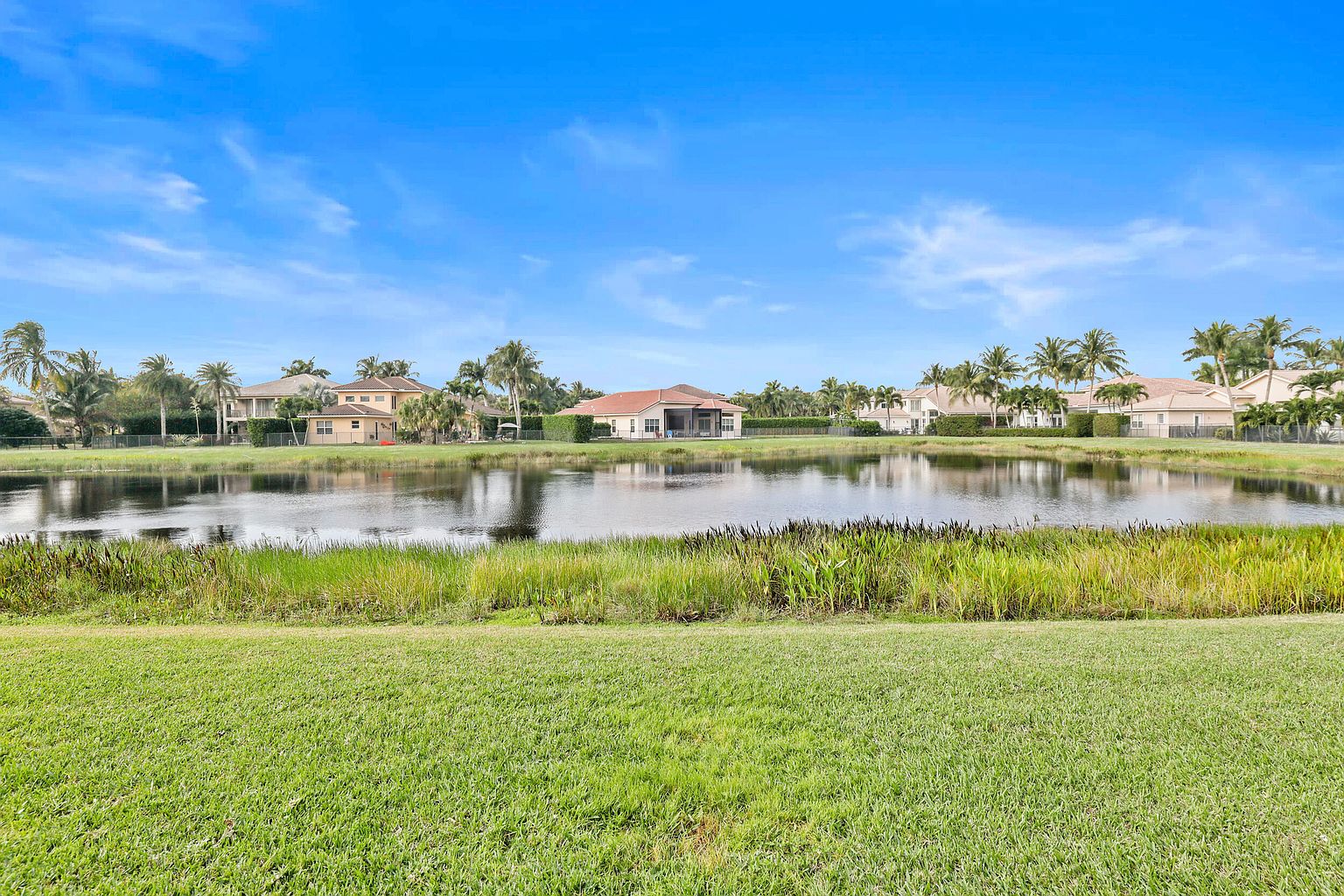 This exterior shot showcases a well-maintained yard with lush green grass leading to a serene pond. Several houses are visible in the background, framed by palm trees, creating a picturesque residential community. The clear blue sky enhances the overall appeal, suggesting a tranquil and desirable living environment.