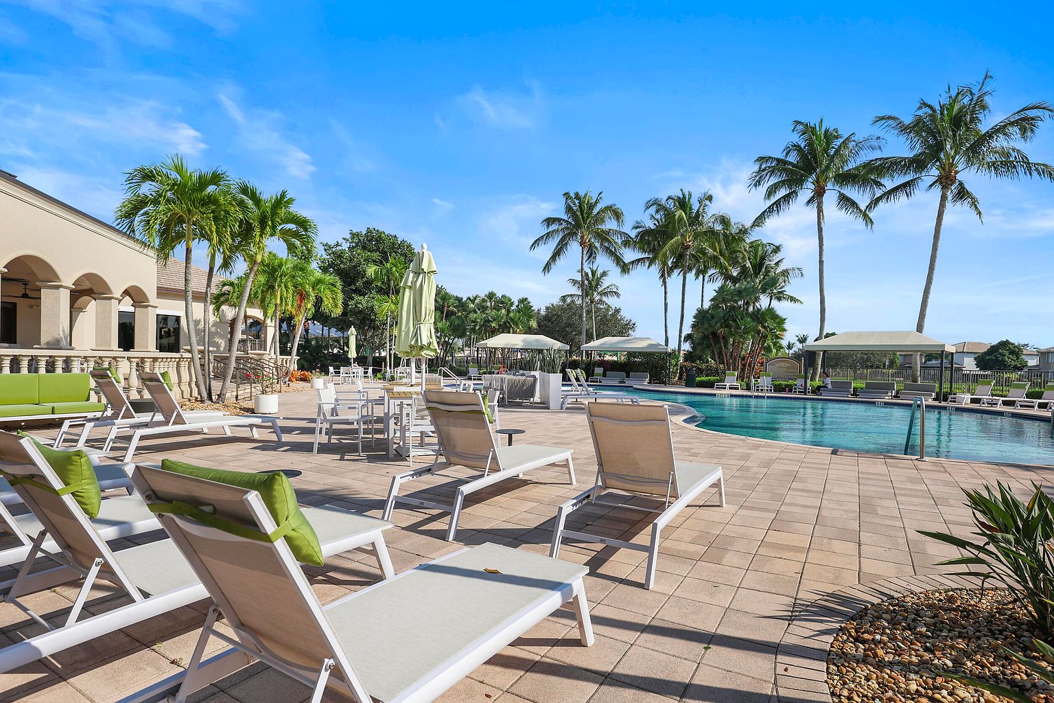This image showcases a luxurious outdoor pool area, complete with lounge chairs, tables, and umbrellas, creating an inviting space for relaxation. The pool is surrounded by lush palm trees and well-maintained landscaping, enhancing the tropical ambiance. The overall impression is one of resort-style living and leisure.