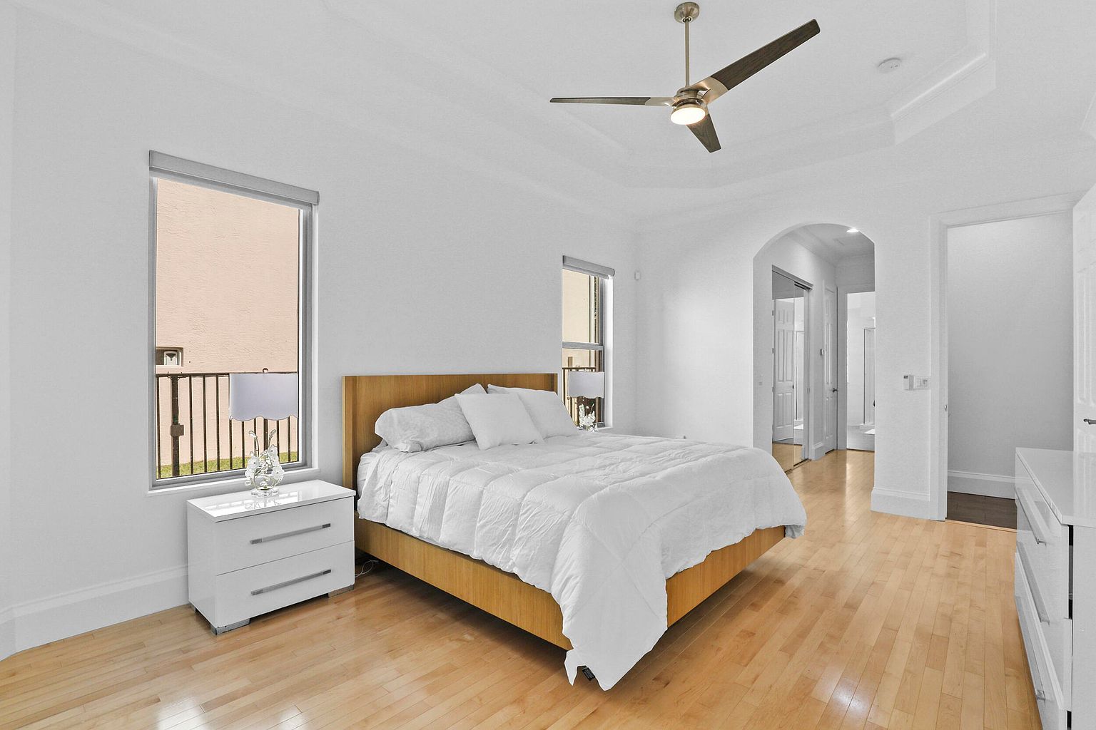 This is a bright and airy primary bedroom featuring a large bed with white linens, light wood flooring, and white walls. Natural light streams in through a window, complemented by a modern ceiling fan. The room has a clean and minimalist aesthetic, creating a serene and inviting atmosphere.