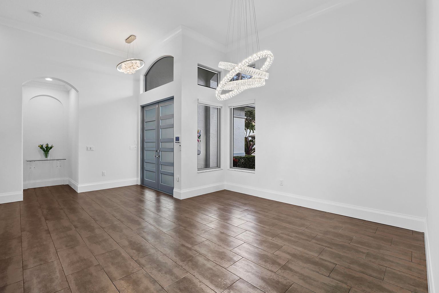 This is an interior shot of a living room, showcasing a modern and spacious design. The room features light-colored walls, dark wood-look tile flooring, and contemporary lighting fixtures. A set of windows provides natural light, and the overall impression is clean and inviting, suitable for entertaining or relaxing.