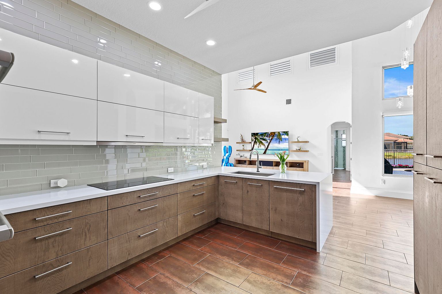 This is a bright, modern kitchen with a combination of white and wood-grain cabinetry, complemented by a white countertop and a light gray backsplash. The kitchen features stainless steel appliances and hardware, and the flooring is a warm-toned wood-look tile. The open layout leads into a living area, creating a spacious and inviting atmosphere.