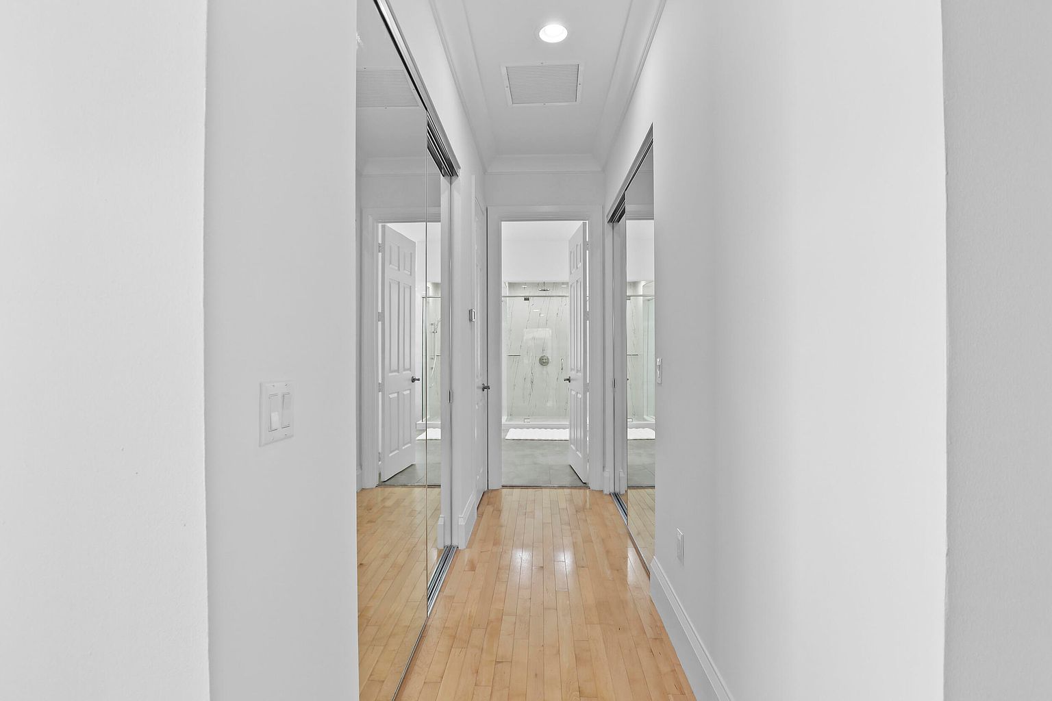 This is an interior shot of a hallway featuring light hardwood floors and white walls. Mirrored closet doors line both sides of the hallway, reflecting the doors to a bathroom with a glass-enclosed shower at the end of the hall. The space is well-lit, creating a clean and modern aesthetic.