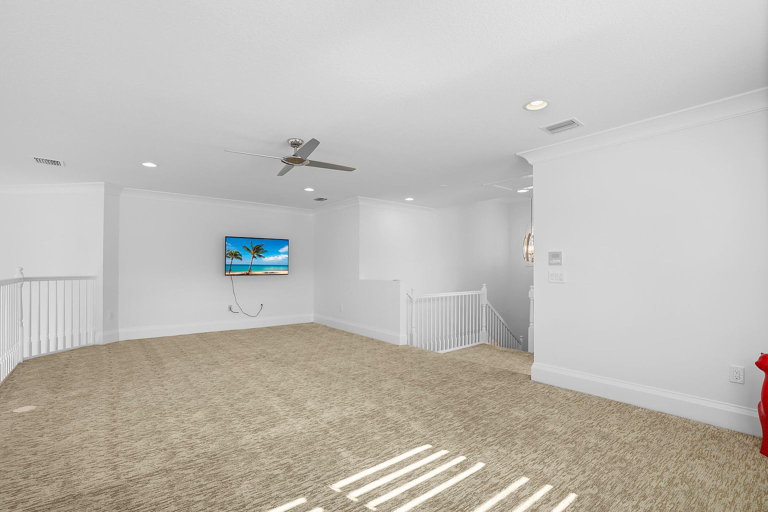 This is an interior shot of a spacious living room featuring neutral-toned carpet and white walls. A flat-screen TV is mounted on the wall, and a ceiling fan provides air circulation. The room has a bright and airy feel, enhanced by natural light streaming through unseen windows, and the staircase adds architectural interest.
