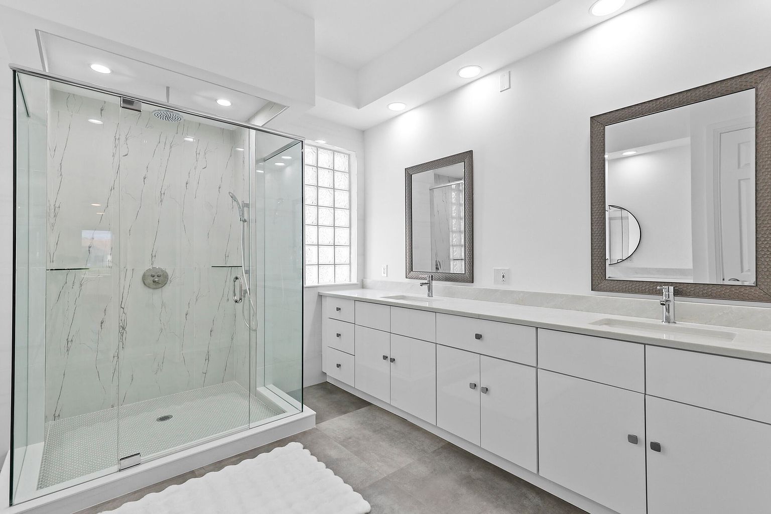 This is a bright and modern primary bathroom featuring a glass-enclosed shower with white marble-style walls and a white tiled floor. The bathroom also has a long white vanity with double sinks, framed mirrors, and gray flooring. The overall impression is clean, spacious, and luxurious.