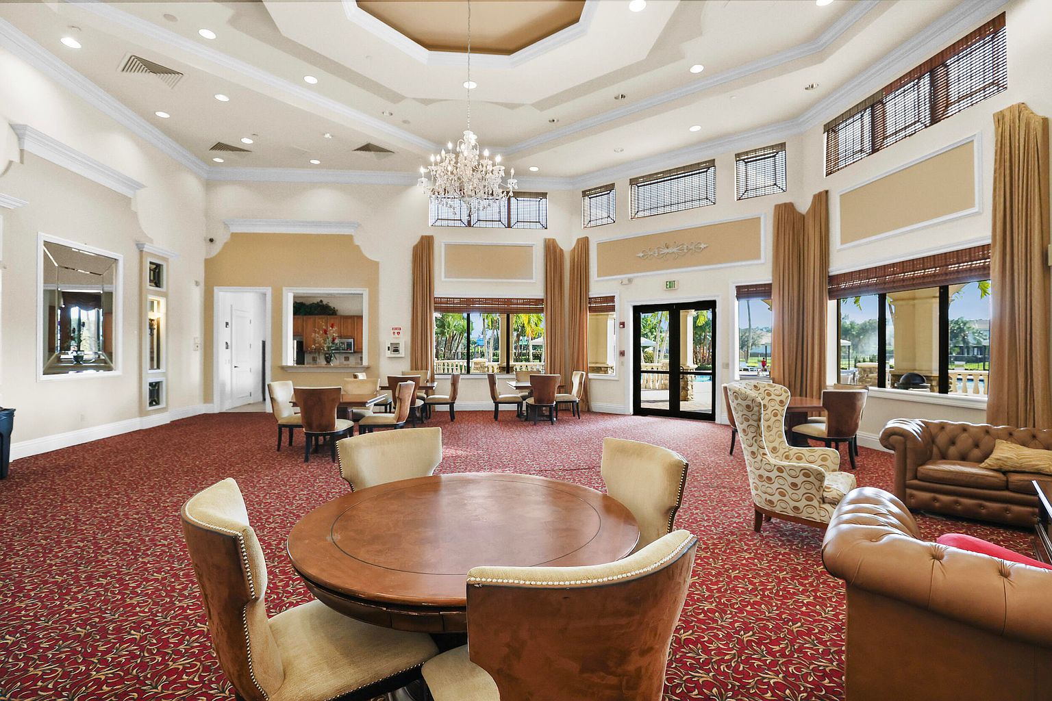 This is an interior shot of a clubhouse amenity space, featuring a large room with multiple seating areas, including round tables with chairs and comfortable sofas. The room is well-lit by a chandelier and natural light from the windows, which are adorned with curtains and blinds. The red patterned carpet adds warmth to the space, creating an inviting atmosphere for residents.