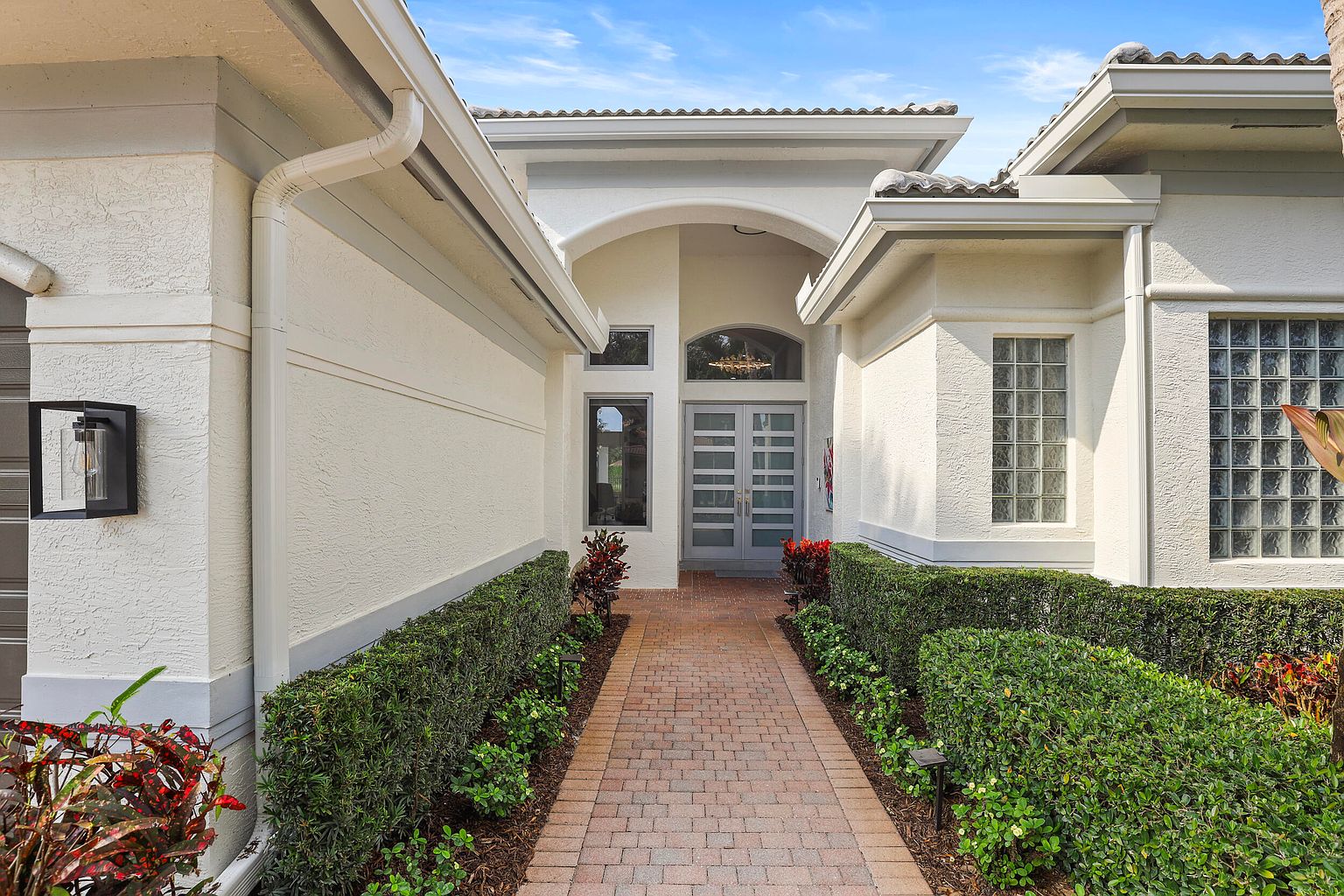 This is an inviting entryway to a home, featuring a brick pathway lined with manicured green hedges leading to a double-door entrance. The house is painted in a neutral tone, complemented by gray accents on the trim and roof. The presence of well-maintained landscaping adds to the curb appeal, creating a welcoming first impression.