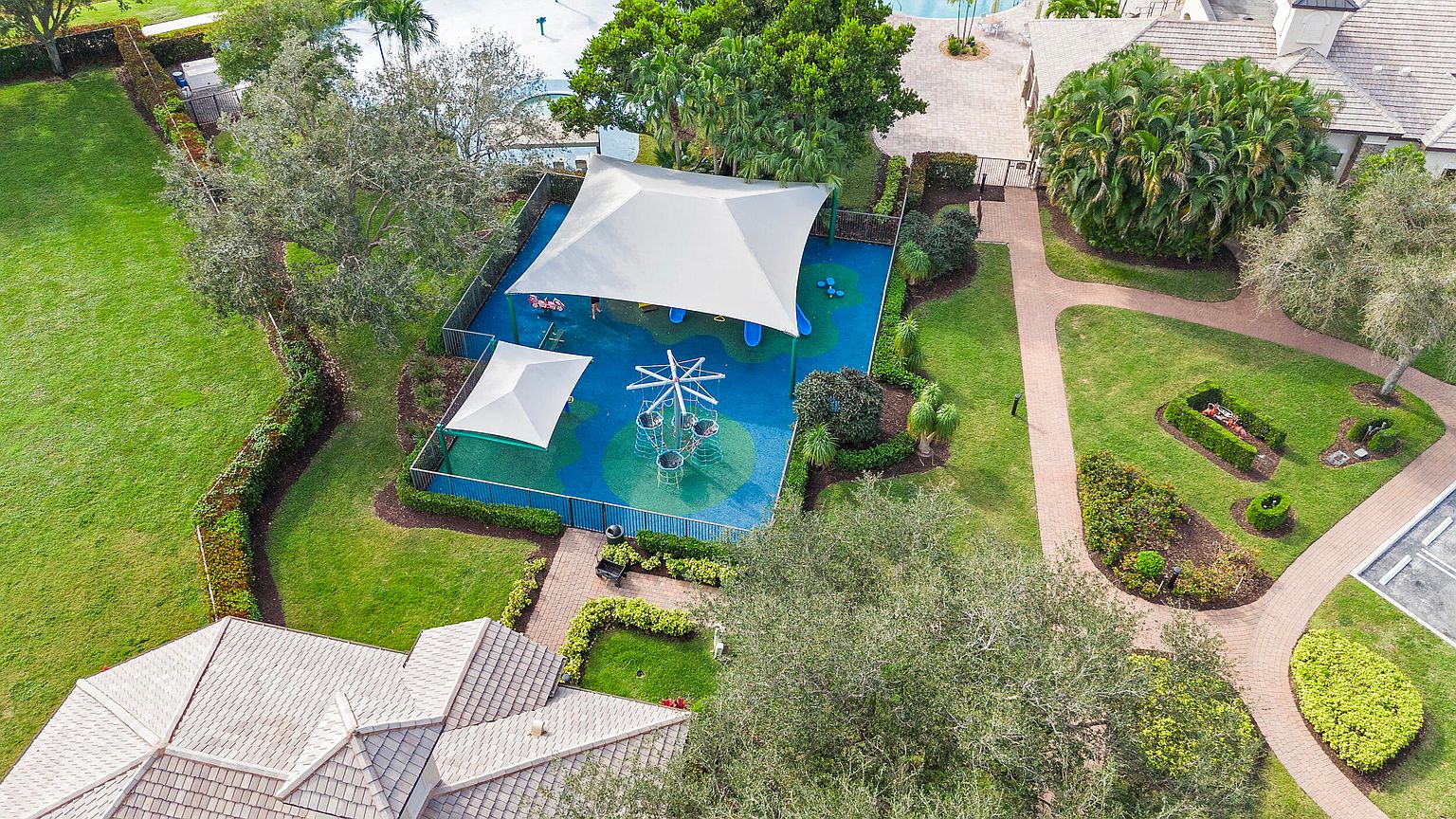 This aerial shot showcases a well-maintained community playground with modern play structures under shade canopies. The playground features a colorful, cushioned surface and is surrounded by lush landscaping and manicured lawns. A nearby brick pathway leads to other community amenities, suggesting a family-friendly environment.