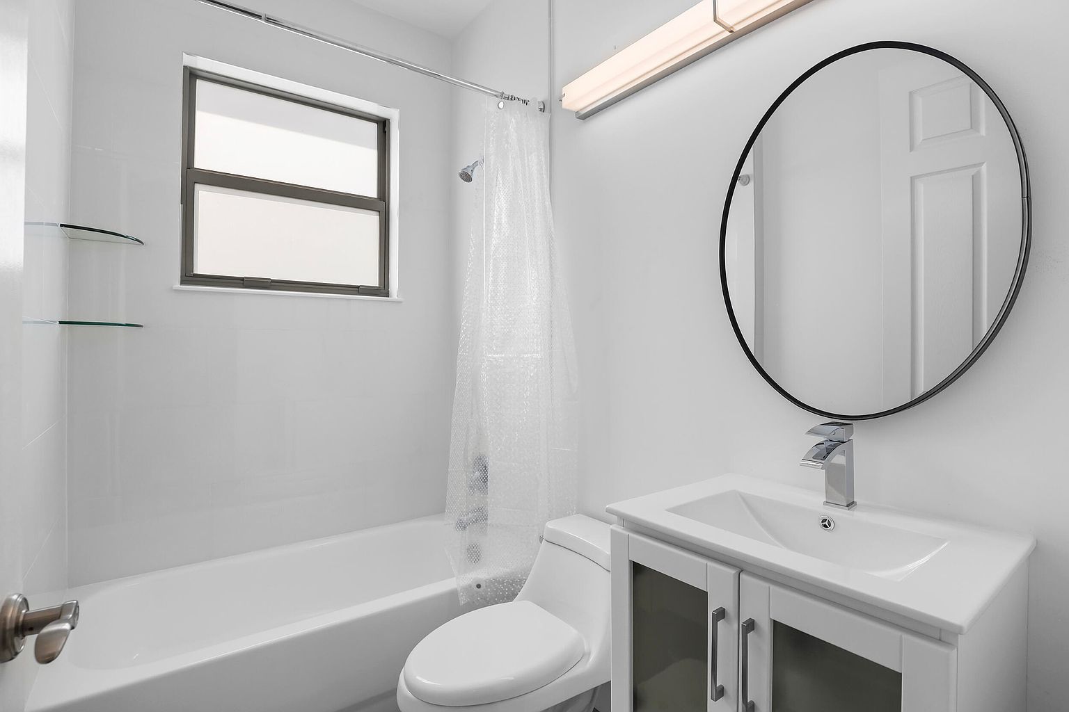This is a well-lit bathroom featuring a white bathtub with a shower curtain, a toilet, and a vanity with a sink and cabinet. A round mirror hangs above the sink, and a light fixture is mounted above the mirror. The bathroom has a clean and modern aesthetic, with white tiles and walls creating a bright and airy space.