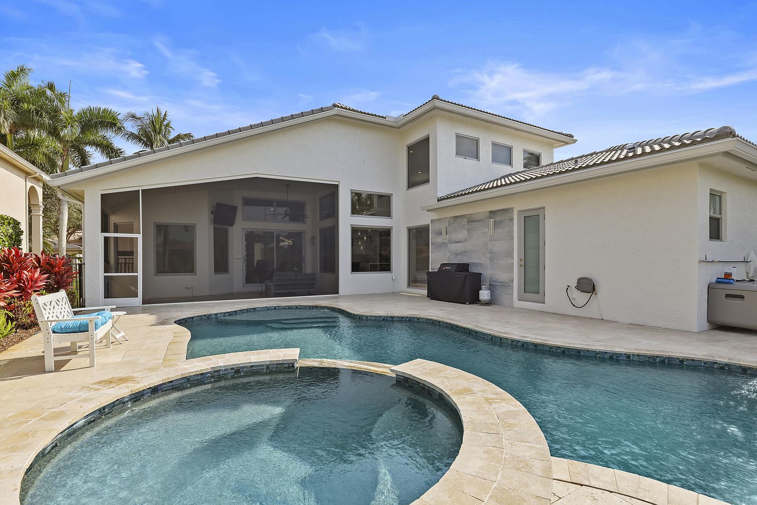 This image showcases a luxurious backyard pool and spa area. The pool features a unique design with a connected spa, surrounded by light-colored stone paving. The house is visible in the background, featuring a screened-in patio and modern architectural elements, suggesting a high-end property perfect for relaxation and entertainment.